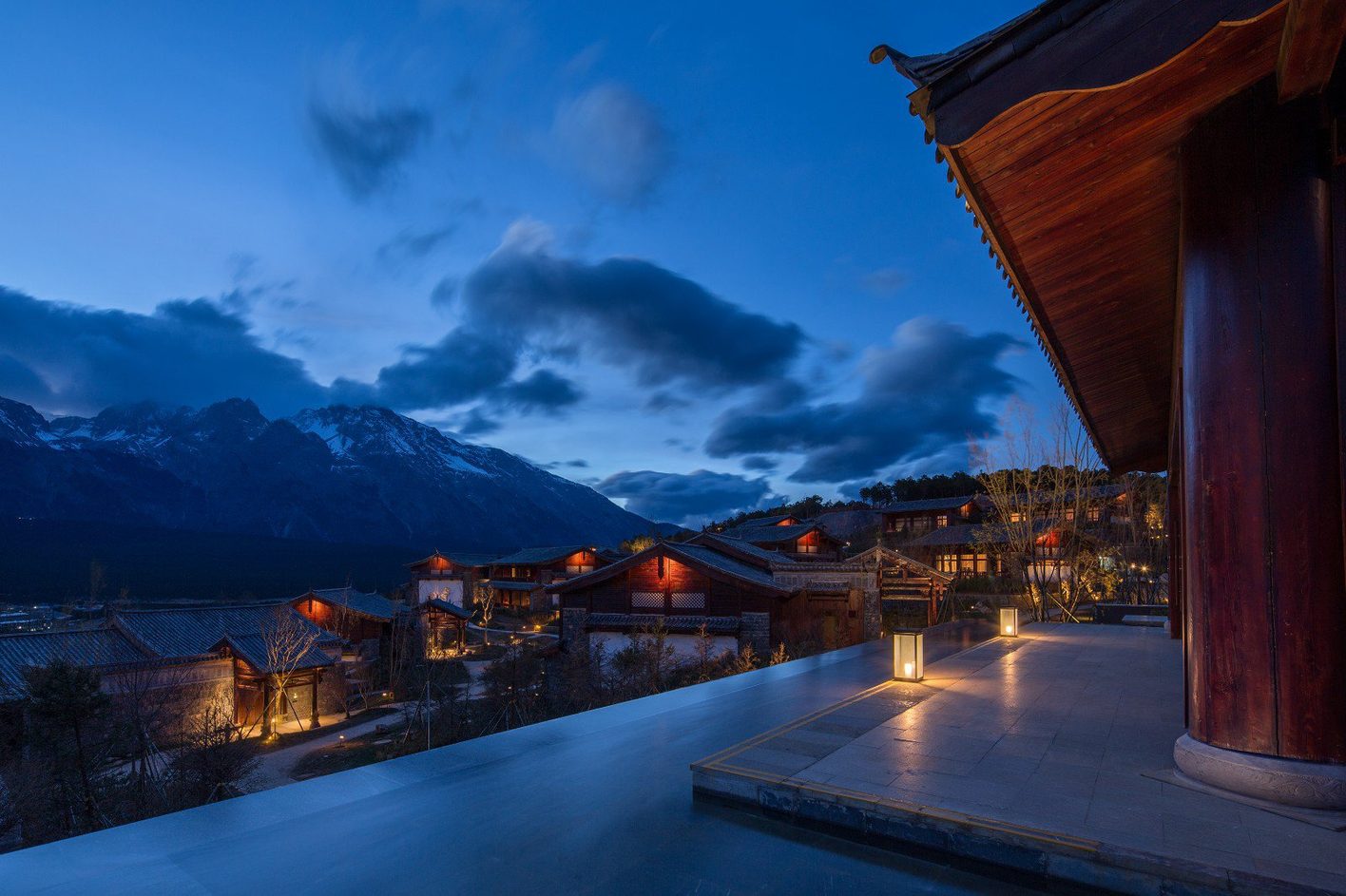 Grand Hyatt Lijiang, Snow Mountain Garden, at dusk: timber village-style buildings under Jade Dragon Snow Mountain, warm light under the eaves and inside, facades almost unlit, floor lanterns on the terrace